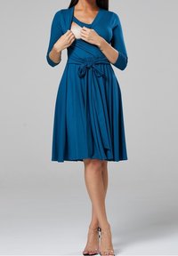 Kvinna i en blå knälång klänning med trekvartsärmar drar åt sidan halsringningen för att avslöja en amningsvänlig öppning.