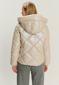 Giubbotto imbottito di un beige chiaro, con una finitura lucida, motivo a rombi e un ampio cappuccio. Indossato con jeans grigi.