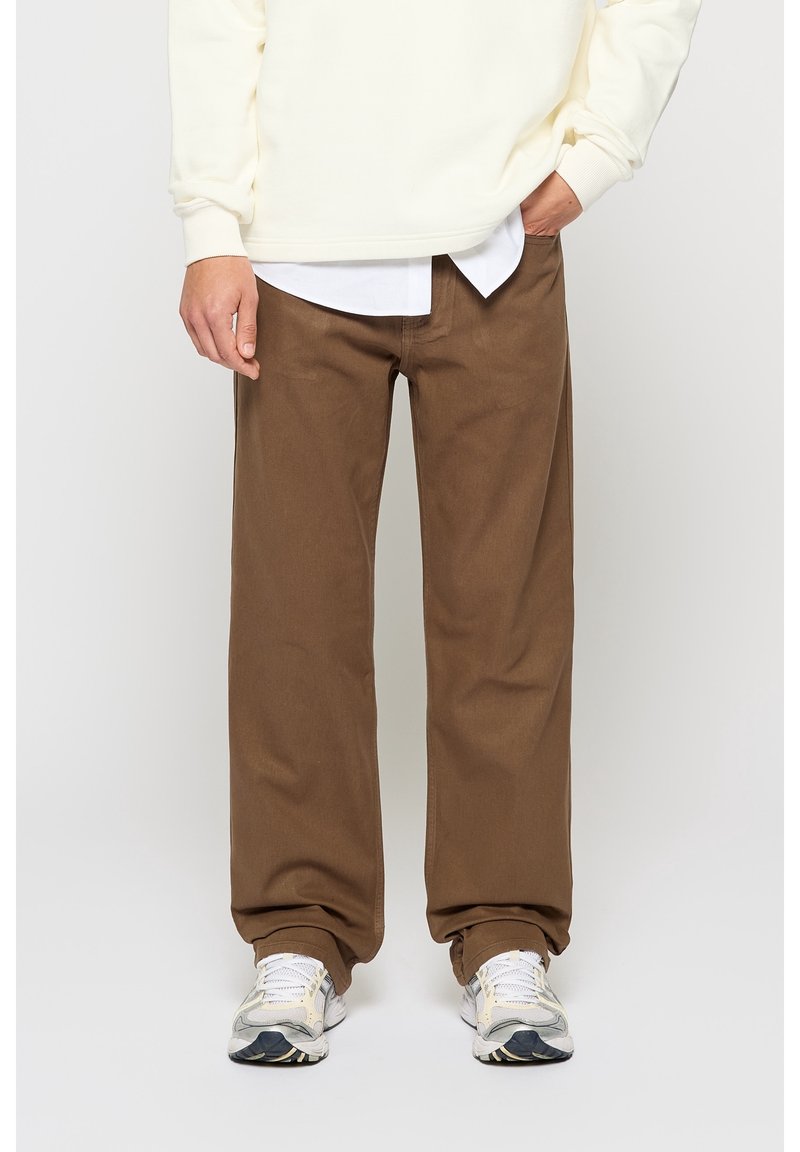 Homme portant un pantalon marron ample, des baskets blanches, une chemise blanche et un pull couleur crème, debout devant un fond uni.