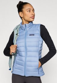 Ανοιχτό γαλάζιο, μονωμένο αμάνικο μπουφάν με ψηλό γιακά, κλείσιμο με φερμουάρ και πλαϊνές τσέπες. Ομαλή υφή, με λογότυπο Patagonia στο στήθος.