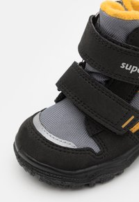 Stivale per bambini nero e grigio con cinturini in velcro, tomaia testurizzata e un rivestimento in pile giallo. Presenta un accento argentato e suola in gomma.