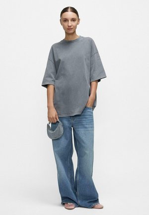 Kobieta stojąca, ubrana w luźną szarą oversize'ową koszulkę, szerokie niebieskie dżinsy i różowe buty, trzymająca w jednej ręce małą szarą torebkę.