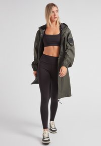Chaqueta impermeable verde oliva con capucha, sujetador deportivo negro y leggings negros. Zapatillas blancas y negras con cordones.
