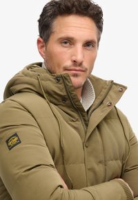 Chaqueta acolchada verde olivo con capucha, que presenta un cuello texturizado, cremallera frontal y una parche de marca en la manga izquierda.