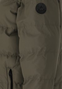 Olivengrøn puffet jakke med ribbet tekstur, lynlåslomme og et sort rundt logo-patch på overarmen.