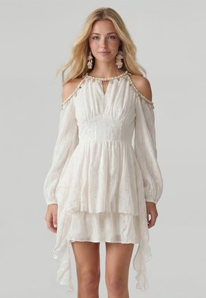 Blonde Frau trägt ein weißes Cold-Shoulder-Kleid mit langen Ärmeln, mehrstufigem Rock und detailliertem Ausschnitt mit hängenden Perlen.