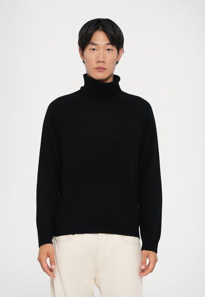 Maglione a collo alto nero con maniche lunghe, realizzato in tessuto morbido, vestibilità comoda e design semplice, indossato sopra pantaloni chiari.
