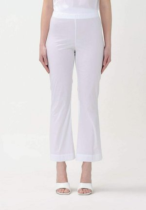 Donna che indossa pantaloni bianchi a gamba dritta e sandali bianchi con tacco e punta aperta, in piedi su uno sfondo bianco uniforme.