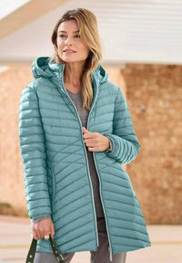 Teal pufferjack met een capuchon, voorzien van een gewatteerd patroon, ritssluiting en een grijze binnenvoering, gecombineerd met een grijze top en denim.