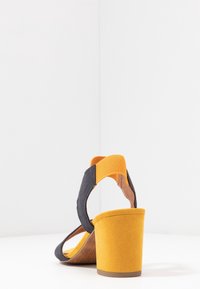 Kaksivärinen sandaali, jossa on tummansininen mokkanahka yläosa, keltainen mokkanahka korko ja joustavat narut. Loivan korkean mallinen ja teksturoitu pohja mukavuutta varten.
