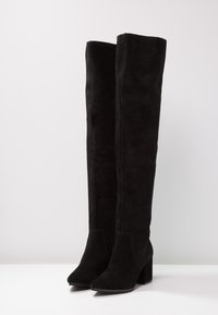 Musta yli polven saappaat, jotka on valmistettu pehmeästä mokasta. Niissä on kärjessä terävä muoto ja paksu korko, sekä sivuvetoketju helppoa pukemista varten.