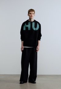 Νέος άνδρας που στέκεται φορώντας μαύρο φαρδύ πουλόβερ με μεγάλα γράμματα "HU", φαρδιά μαύρα παντελόνια και μαύρα παπούτσια μπροστά σε απλό φόντο.