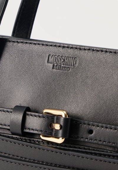 Détail d'un sac à main Moschino Milano en cuir noir montrant le logo de la marque cousu et une boucle dorée sur la sangle réglable.