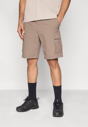 Shorts cargo beige avec une poche latérale, en tissu léger. Associés à des chaussures de sport noires et des chaussettes noires côtelées.