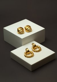 Goldene Ohrringe auf weißen Ständern ausgestellt: runde, gerippte Stecker und ein gedrehter Creolen-Design, beide mit einer glänzenden metallischen Oberfläche.