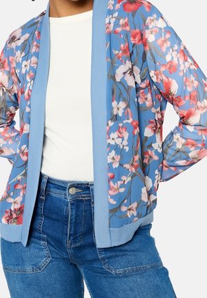 Femme portant une veste bleu clair à motif floral par-dessus un top blanc et un jean bleu, les mains posées sur les hanches.