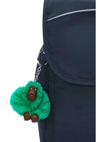 Lo zaino navy presenta un portachiavi a forma di scimmietta verde peluche con un viso marrone, una chiusura lucida e una consistenza soffice. Superficie del tessuto liscia visibile.