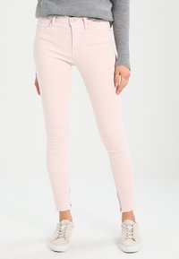 Jeans ajustados en denim rosa claro con una textura suave, cintura de altura media y cremalleras en los tobillos, combinados con zapatillas beige.