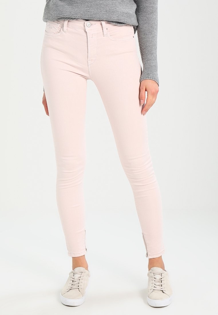 Jeans ajustados en denim rosa claro con una textura suave, cintura de altura media y cremalleras en los tobillos, combinados con zapatillas beige.