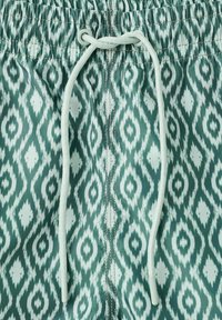 Ceinture élastique avec cordon blanc sur tissu vert présentant un motif géométrique losange blanc.