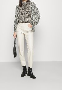 Blouse noire et crème à imprimé floral avec manches bouffantes, associée à un pantalon crème et des bottines noires, tenant un sac noir texturé.