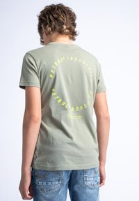 Groene katoenen T-shirt met korte mouwen en een ronde hals. Heeft grote gele tekst op de achterkant, boven de zoom, in een cirkelvormige lay-out.