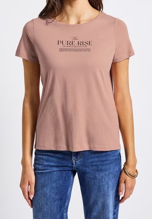 Kobieta w krótkim rękawie w kolorze kurzowej różu z napisem "The Pure Rise", niebieskich dżinsach i złotych bransoletkach na prawym nadgarstku.