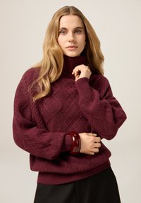 Vinröd stickad polotröja med texturerade diamantmönster, med vida ärmar och ribbad fåll, kombinerad med ett mörkt armband.