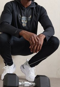 Schwarzes Langarm-Hoodie-Shirt mit grauem Bullenlogo; kombiniert mit schwarzen Leggings und weißen Sportschuhen. Ein Hantel liegt im Vordergrund.