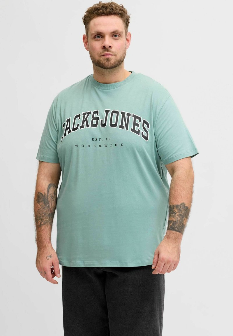 Homme tatoué portant un t-shirt vert menthe Jack & Jones et un pantalon noir, debout devant un fond blanc uni.