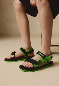 Zwarte en groene kindersandalen met verstelbare banden, een textuurs zool, oranje en groene accenten, en een Minecraft-logo aan de zijkant.