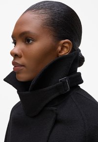 Black wool coat with high collar, featuring a buckle detail on the collar, structured shape, and soft texture.