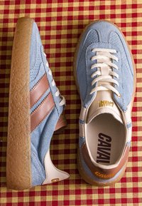 Paire de baskets en daim bleu avec des rayures marron et des semelles en gomme sur une surface en tissu à carreaux rouge et jaune.