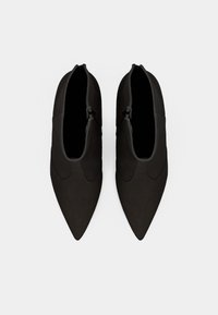 Botas de tornozelo pretas de bico fino com uma textura suave e desenhos curvos subtis nas laterais. Fechadas com um ziper na parte de trás.