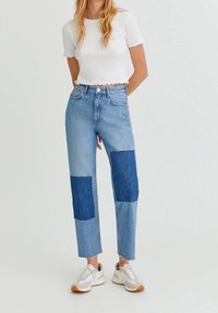 Jean bleu clair avec des morceaux de denim bleu foncé sur le devant, taille haute, coupe droite ; porté avec un top court blanc et des baskets.