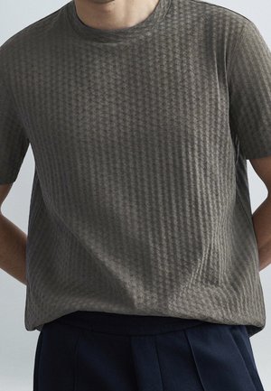 Mężczyzna ubrany w szary, teksturowany t-shirt z krótkim rękawem i ciemnoniebieskie spodnie na jednolitym tle.