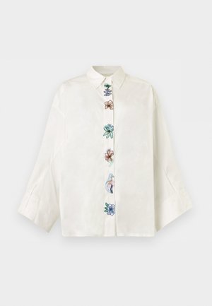 Kvinne iført en overdimensjonert hvit bluse med blomsterbroderi foran, lyseblå vide jeans og svarte spisse sko.