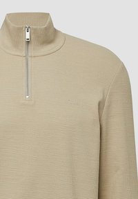 Beżowy sweter zapinany na zamek błyskawiczny z wysokim kołnierzem, teksturowana tkanina, prążkowany wzór oraz małe logo na piersi. Szczegół z zamkiem błyskawicznym w kolorze srebrnym.