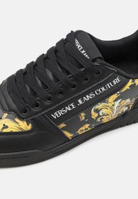 Zwarte sneakers met een glad leren bovenwerk, voorzien van gouden en witte bloemaccenten, zwarte veters en een wit logo-opdruk op de tong.