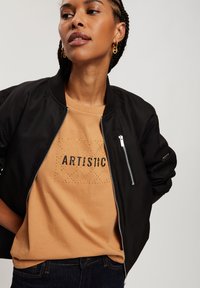 Czarna kurtka bomberka z zamykanymi kieszeniami, noszona na beżowym t-shircie z napisem "ARTISTIC" w zdobionych literach. Ciemne dżinsy dopełniają stylizację.