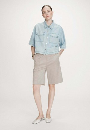 Femme debout les mains dans les poches portant une chemise en jean bleu clair raccourcie, un short long beige, des chaussures blanches à petit talon et des boucles d'oreilles.