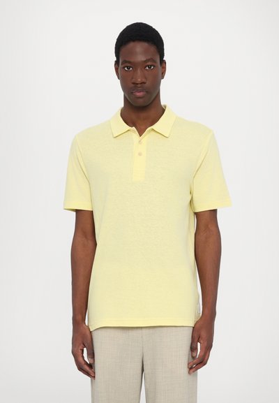 Polo de manga corta en suave tela amarilla, con un panel de tres botones y una sutil textura. Combinado con pantalones gris claro.