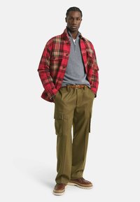 Junger Mann mit geflochtenem Haar trägt eine rot-gestreifte Jacke, einen grauen Half-Zip-Pullover, olivfarbene Cargo-Hosen, einen geflochtenen braunen Gürtel und braune Schuhe.