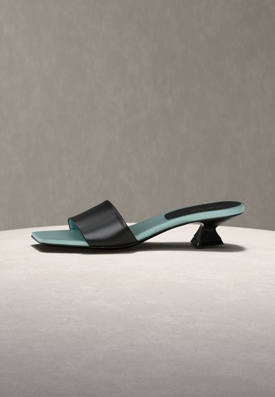 Μαύρο ανοιχτό σανδάλι mule με τετράγωνη μύτη, χαμηλό σκαλιστό τακούνι και ανοιχτό μπλε πάτο, εκτεθειμένο σε ουδέτερη επιφάνεια.