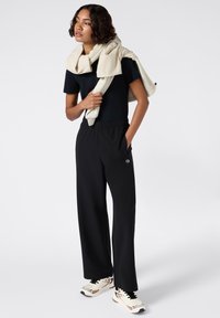 Zwarte relaxed-fit broeken met een elastische tailleband, gecombineerd met een aansluitend zwart topje en een crèmekleurige trui die over de schouders hangt. Sneakers maken de look af.