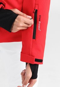 Rød vejrbestandig jakke med sildebensmønster, med en sort lynlåslomme og velcro manchet, mærket "Clima Protect®".