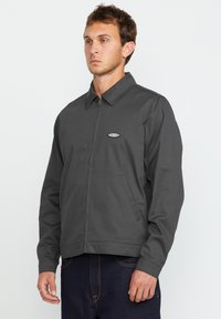 Grijze zip-up jas met een kraag, zijzakken en een logo patch op de borst. Glad materiaal met een iets gestructureerde pasvorm.