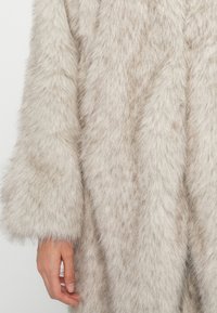 Cappotto in pelliccia sintetica di un beige chiaro con una superficie morbida e testurizzata e maniche ampie. Presenta una vestibilità rilassata con un aspetto lussuoso e soffice.
