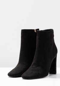 Un par de botas de tobillo de ante negro con tacones cuadrados y cremalleras laterales sobre una superficie blanca y fondo liso.