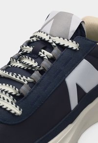 Σκούρο μπλε αθλητικό παπούτσι με παχιές πλεξούδες κορδόνια σε λευκό και γκρι, λευκό πλευρικό λογότυπο και εμφανές μαλακό μπεζ πάτο.